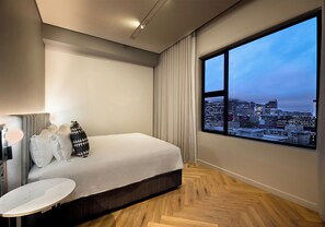 A modern hotel room with a large window offering a city view, a bed with white bedding, a round marble-topped table, and a wall-mounted TV.
