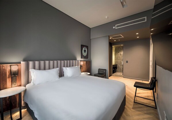 A modern hotel room with a large bed, a small table, a chair, and a bathroom visible through an open door.