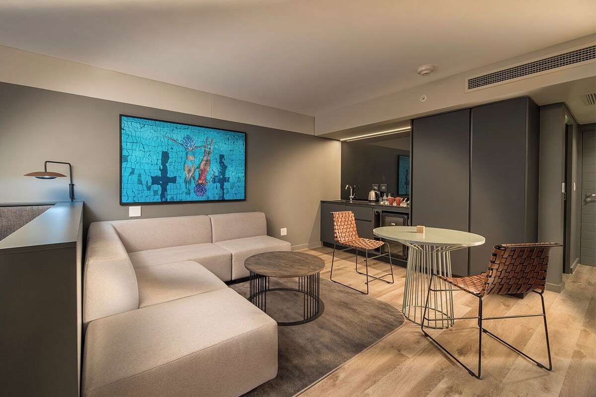 A modern living room with a sofa, round coffee table, and two chairs. There is a kitchen area in the background with a dining table and chairs.