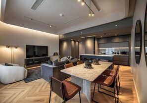 A modern living room with a large TV, comfortable seating, a dining area with a table and chairs, and a kitchen in the background.