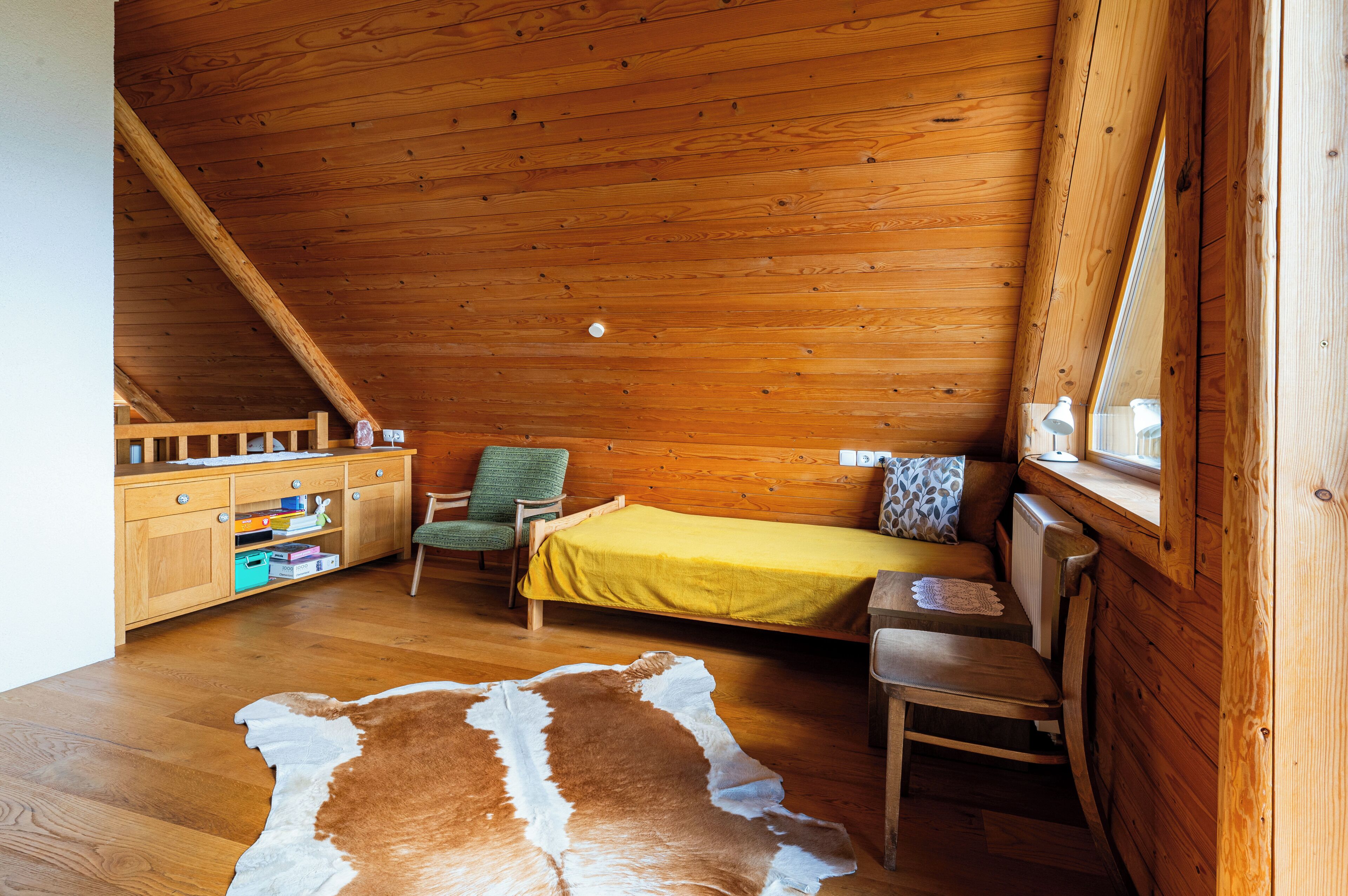 Una habitación de cabaña acogedora con techo de madera, una cama con manta amarilla, una silla verde, un escritorio de madera con espacio de almacenamiento y una alfombra de cuero de vaca.
