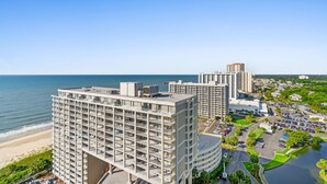 A high-rise hotel with multiple floors, a parking lot, and a beach view.