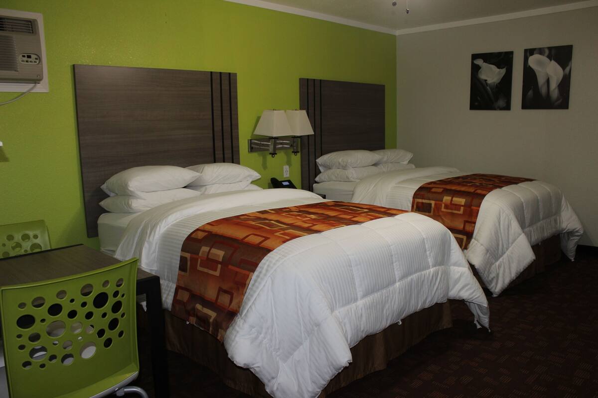 Habitación de hotel con dos camas, una silla verde, un escritorio y una lámpara de pared.