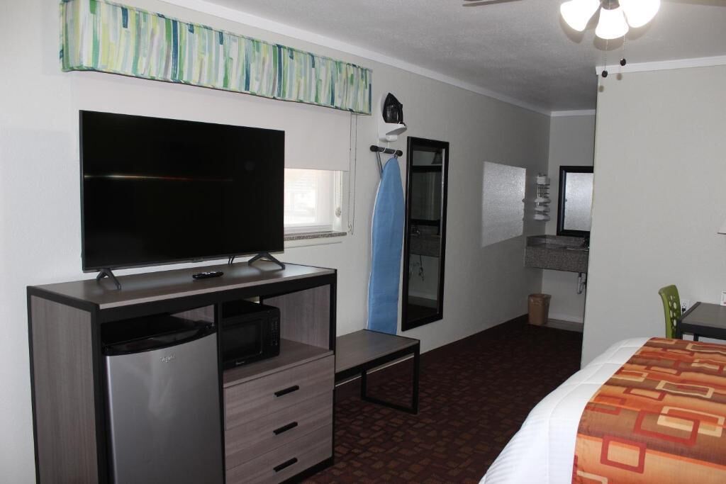 A hotel room with a flat-screen TV, a small desk with drawers, a bed with a patterned bedspread, a ceiling fan, and a window with a curtain.