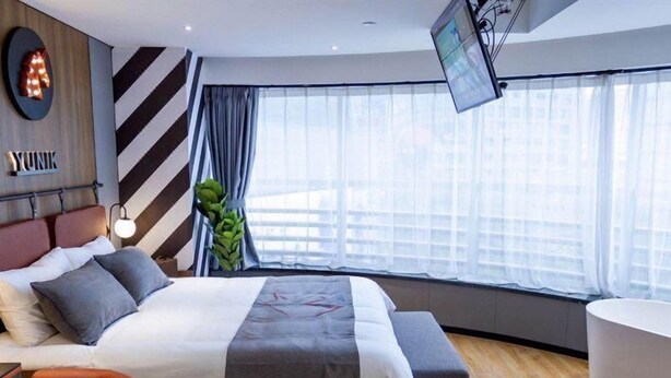 Habitación de hotel moderna con una cama grande, un área de descanso con una butaca, un ventanal con cortinas y un televisor de pantalla plana montado en la pared.