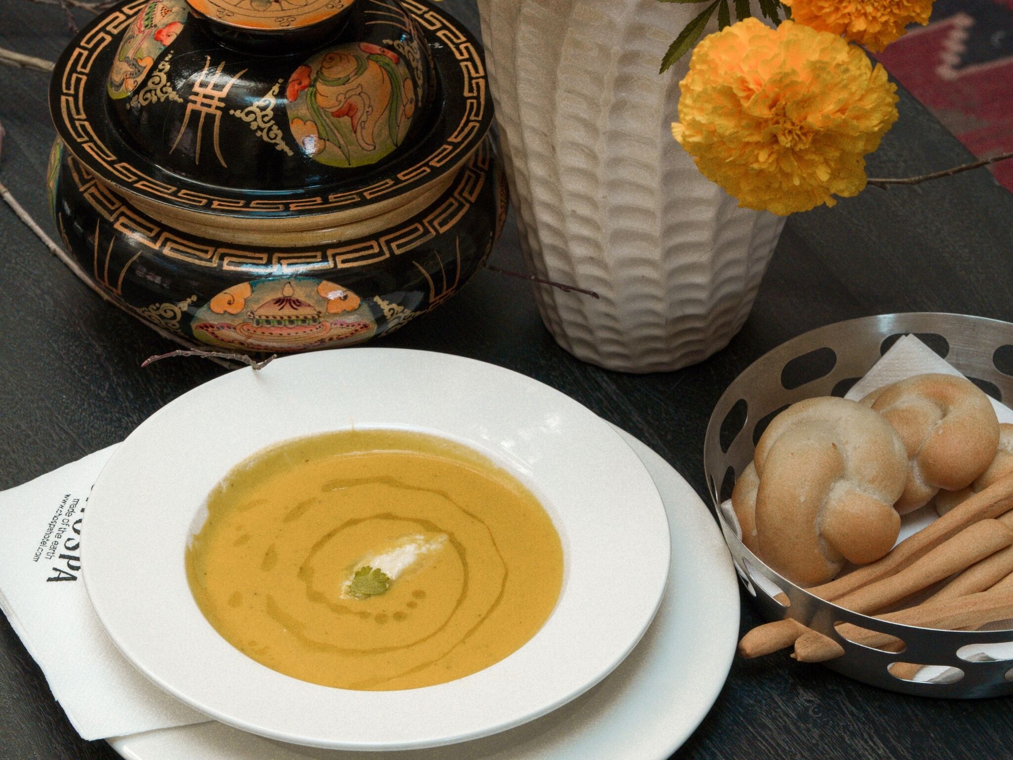 A bowl of soup with a decorative lid, a white vase with flowers, and a basket of breadsticks.