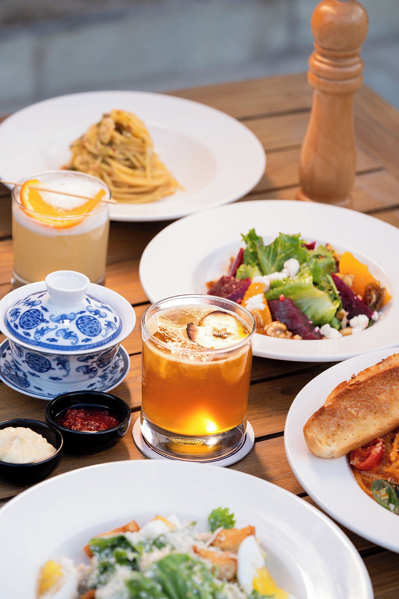 Una mesa de madera con varios platos, incluyendo pasta, ensalada y tostadas, además de un vaso de bebida de color ámbar y un recipiente pequeño de cerámica azul y blanco.