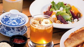 Una mesa de madera con varios platos, incluyendo pasta, ensalada y tostadas, además de un vaso de bebida de color ámbar y un recipiente pequeño de cerámica azul y blanco.