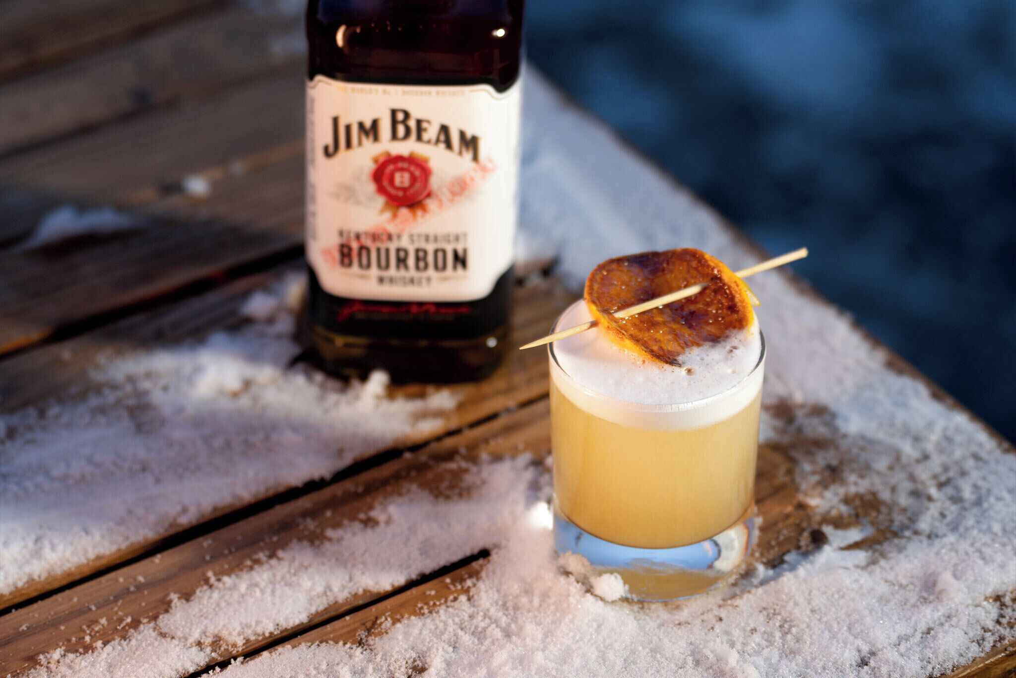 Una botella de Jim Beam Bourbon y un vaso con un cóctel sobre una mesa de madera cubierta de nieve.