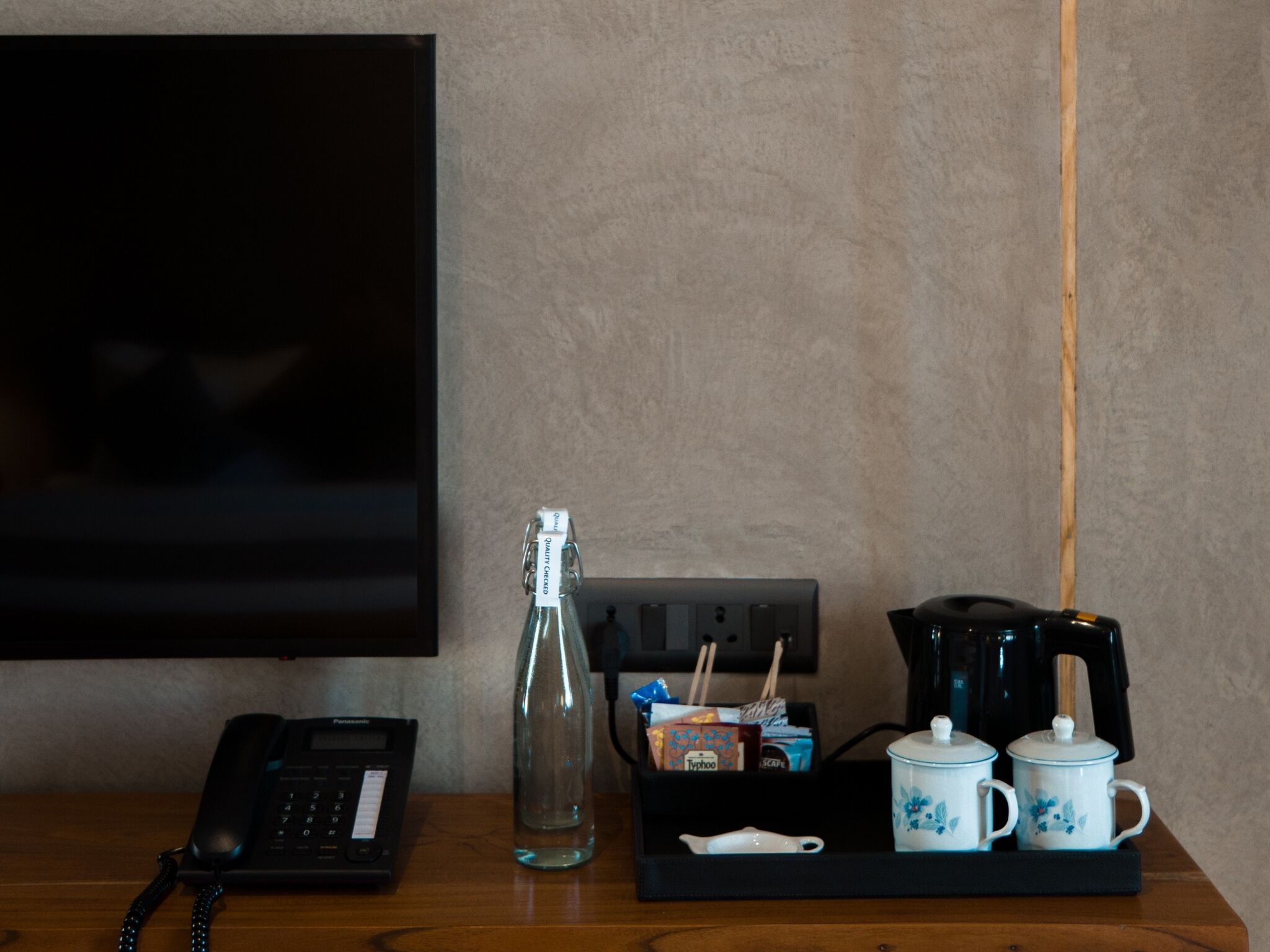 Habitación de hotel con un escritorio de madera, un teléfono negro, una botella de vidrio, una cafetera y dos tazas florales sobre una bandeja.