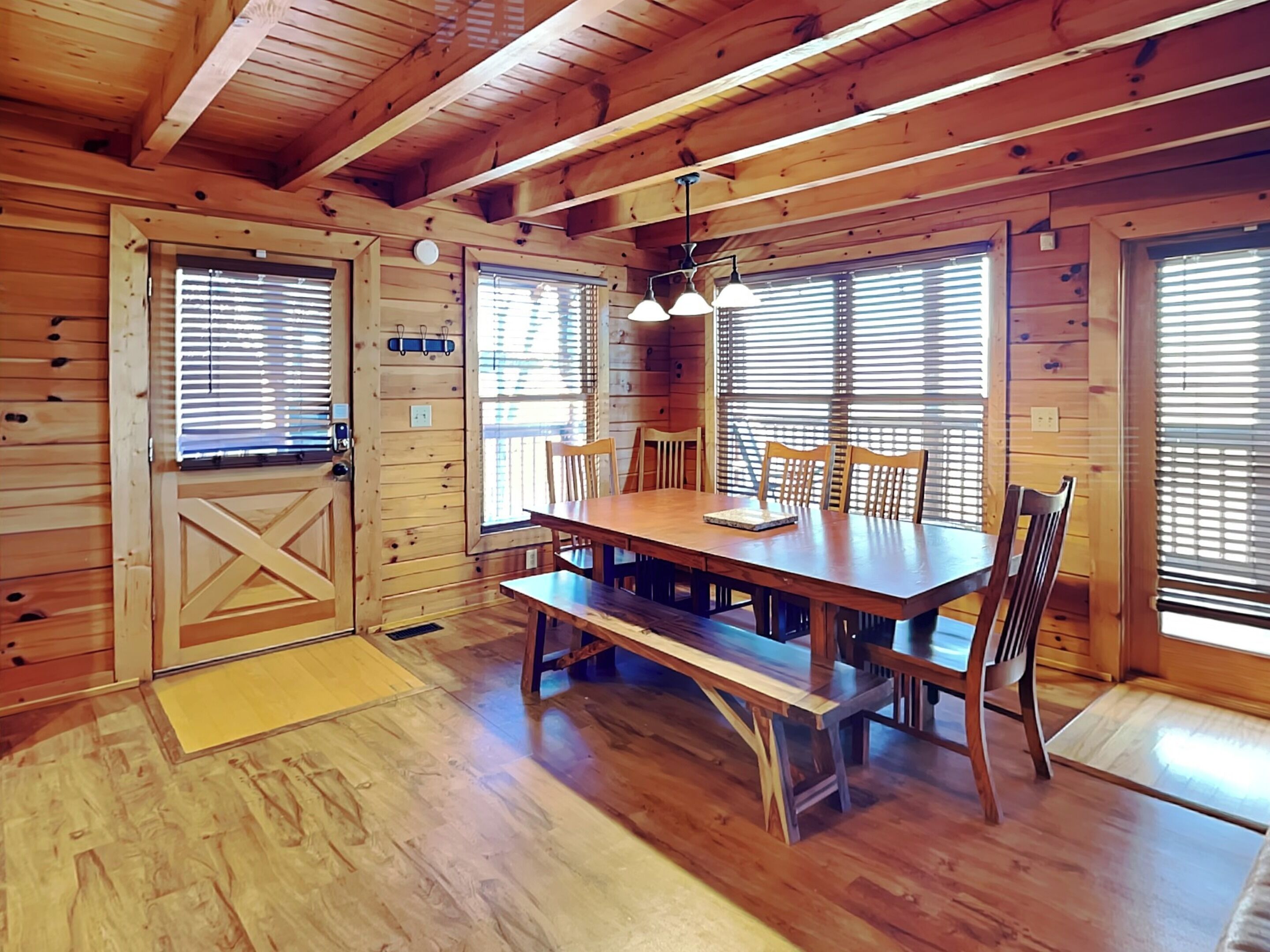 A wooden cabin with a dining table and chairs, large windows with blinds, and a ceiling with exposed beams.