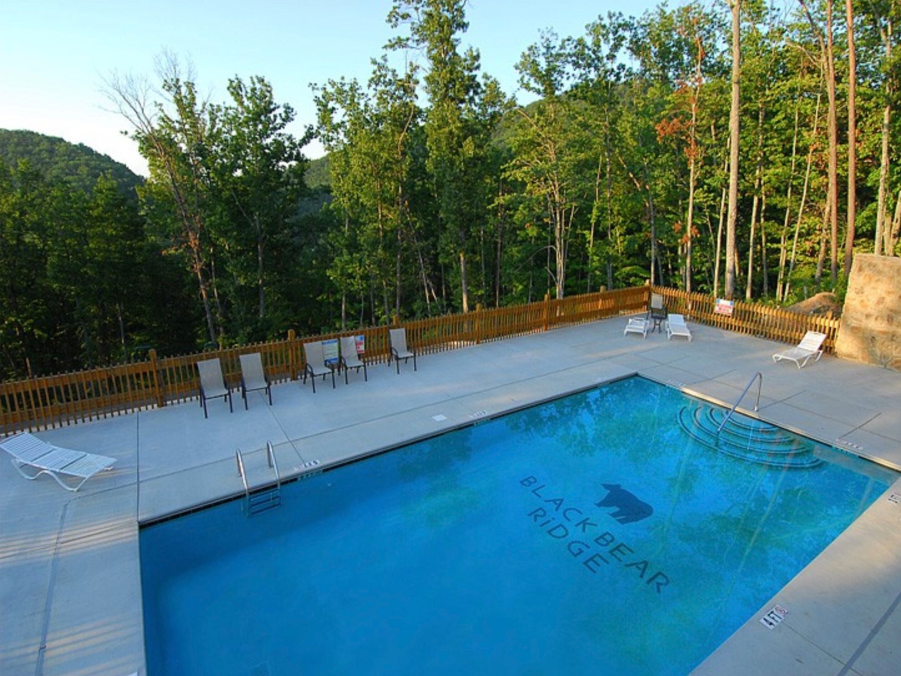 En rektangulär swimmingpool med texten ”BLACK BEAR RIDGE” målad längs kanten, omgiven av ett trästaket, och med träd i bakgrunden.