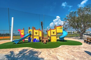 A playground with slides and climbing structures, surrounded by a fence and a tree. - Madeira Beach Yacht Club 151H by Distinctive Beach Rentals (Madeira Beach)
