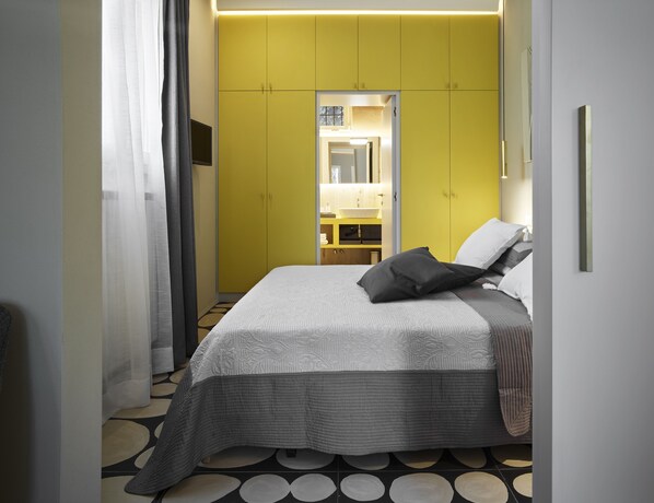 Une chambre à coucher moderne avec un lit, une armoire jaune, un miroir et une salle de bain visible à travers une porte ouverte.