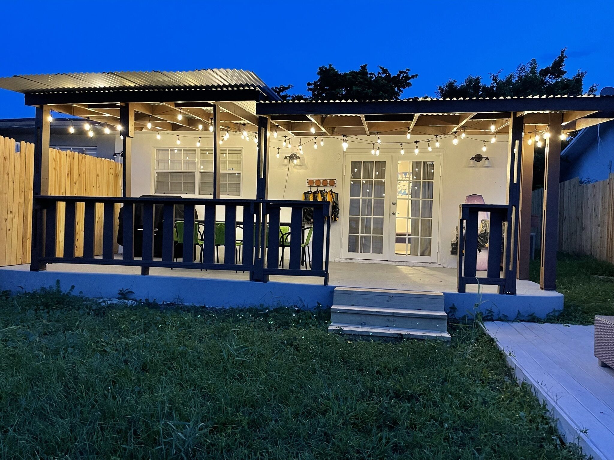 A covered outdoor area with a wooden deck, string lights, and a glass door leading to a building.