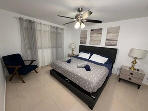 A bedroom with a bed, a chair, a nightstand, a lamp, a ceiling fan, and curtains.