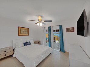 Quarto com cama, ventoinha no teto, mesa de cabeceira, cômoda, televisão e janela com cortinas.