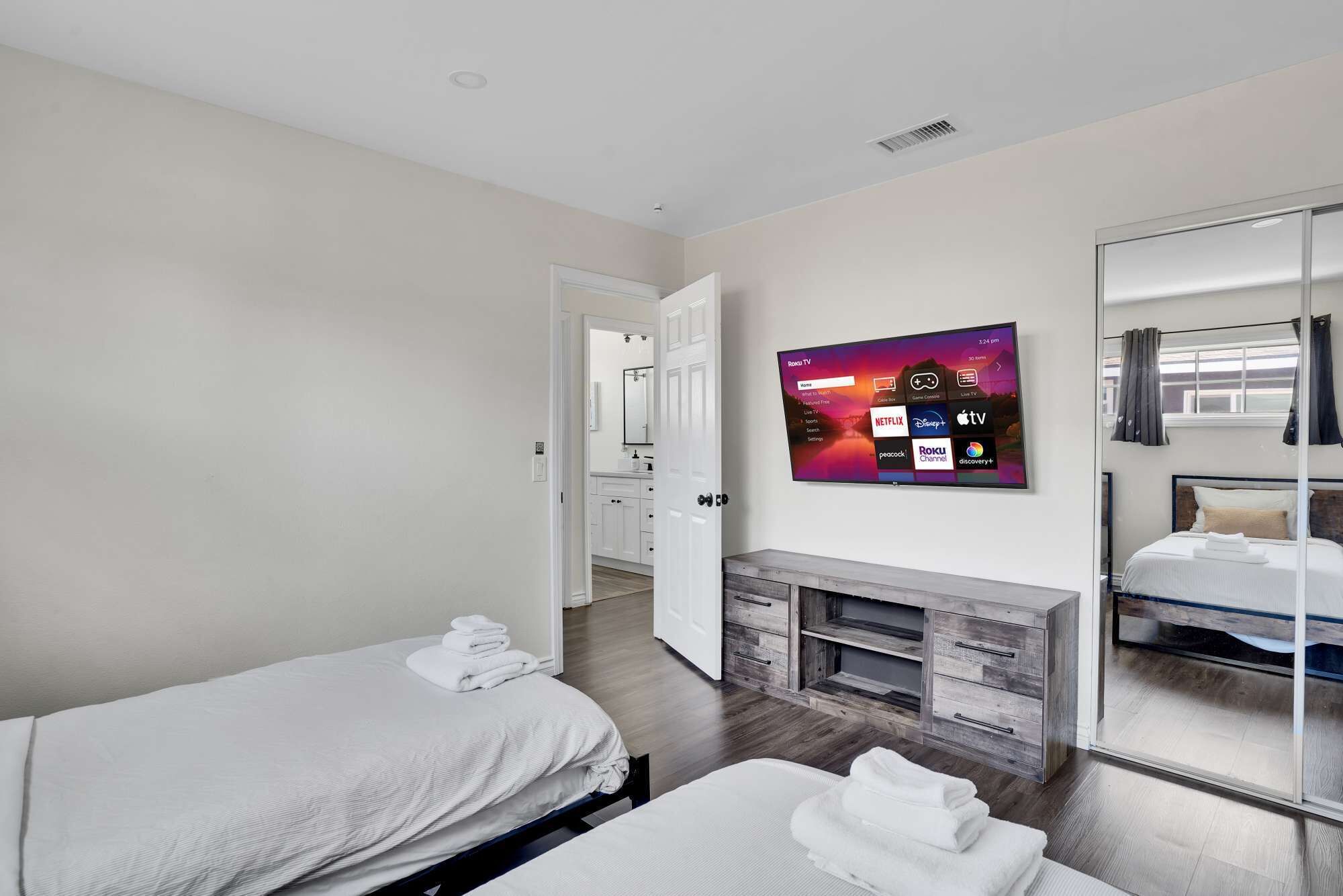 A hotel room with two beds, a TV mounted on the wall displaying various streaming services, a wooden console table, and a large mirror.
