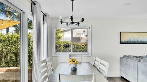 A dining area with a table, chairs, and a chandelier. There is a window with blinds and a view of greenery outside.