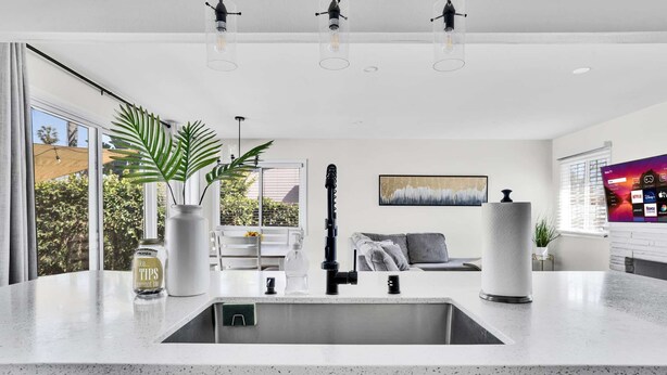 A modern kitchen with a large island, a built-in sink, and pendant lighting. There is a potted plant and a painting on the wall.