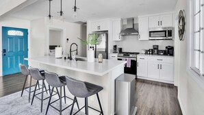 A modern kitchen with a central island, bar stools, white cabinets, stainless steel appliances, and a blue door.