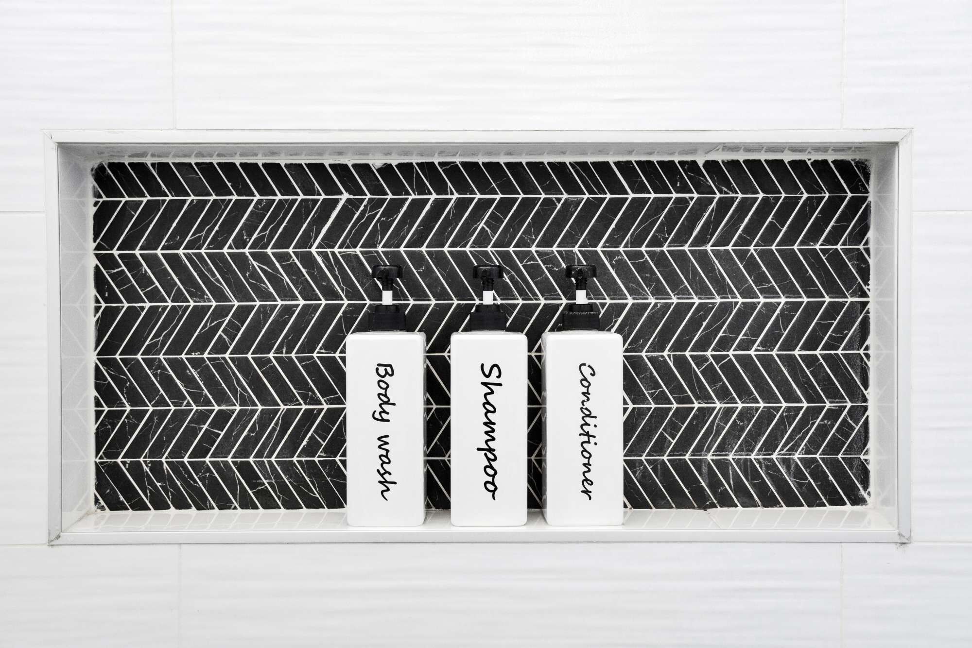 Three white bottles labeled "Body wash," "Shampoo," and "Conditioner" in a window with a black and white herringbone pattern backsplash.