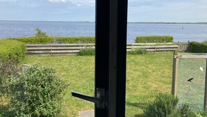 Une vue à travers une fenêtre sur une zone herbeuse, avec une clôture en bois, une étendue d’eau et une rive lointaine.