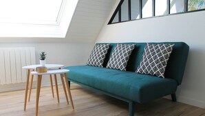 A teal sofa with patterned cushions, a small table with a plant, and a white radiator in a room with wooden flooring and a skylight. - Cabourg - Cosy studio 1 min from the beach (Cabourg)