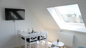 A room with a white table and chairs, a small round table with a plant, a TV mounted on the wall, and a skylight. - Cabourg - Cosy studio 1 min from the beach (Cabourg)