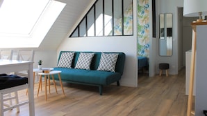 A loft-style room with a sloped ceiling, a teal sofa with patterned cushions, a dining table with chairs, a small stool, and a mirror on the wall. - Cabourg - Cosy studio 1 min from the beach (Cabourg)