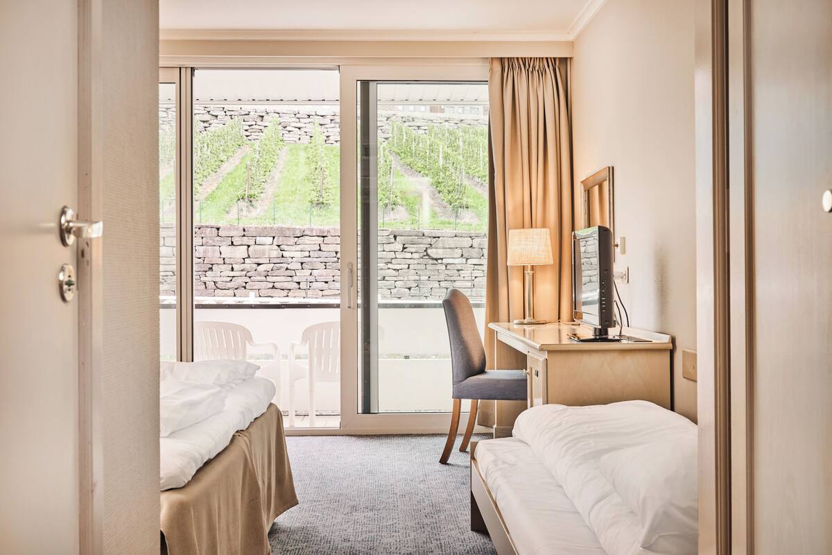 A hotel room with a bed, a desk, a chair, and a large window with a view of a stone wall and greenery.