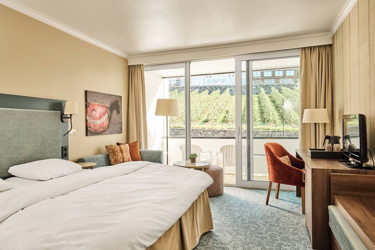 A hotel room with a large bed, a desk, a chair, and a balcony with a view of the outdoors.
