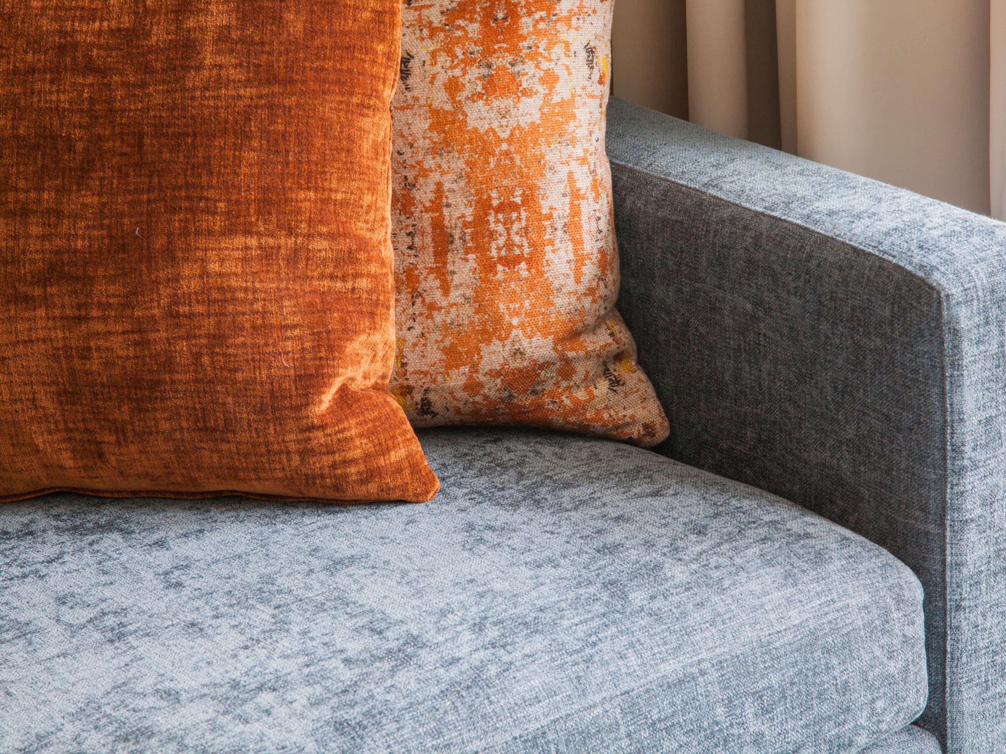 A grey upholstered sofa with two pillows, one orange and one white with orange spots.