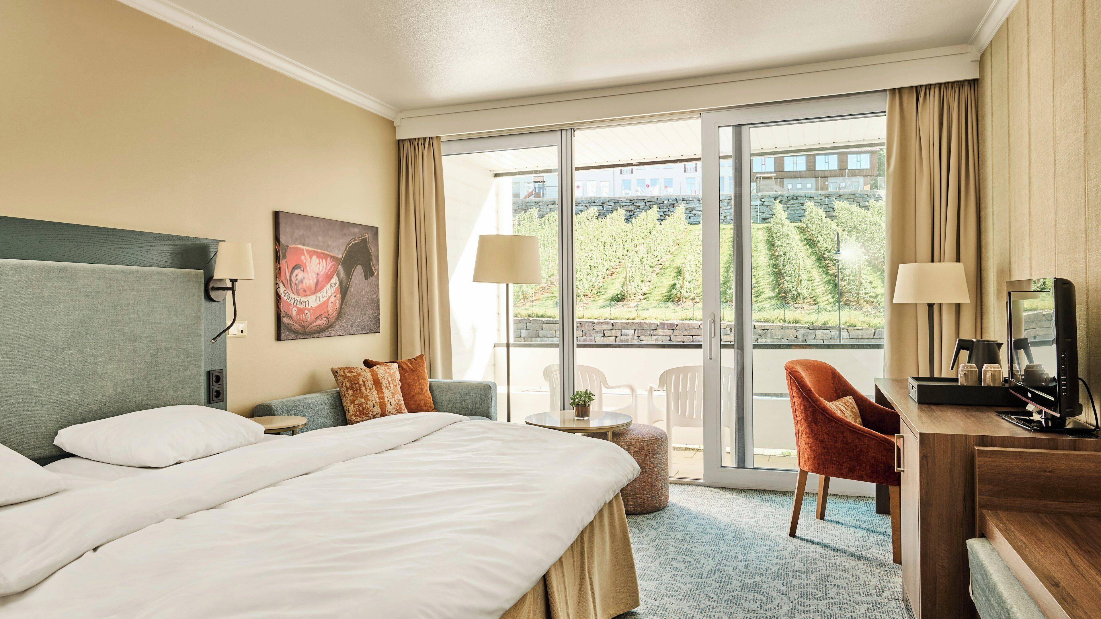 A hotel room with a large bed, a desk with a computer, a chair, a small table with a plant, and a balcony with a view of greenery.