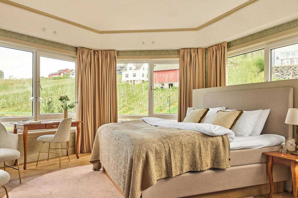 A bedroom with a large bed, a desk, and a chair, a window with curtains, and a view of greenery and buildings.