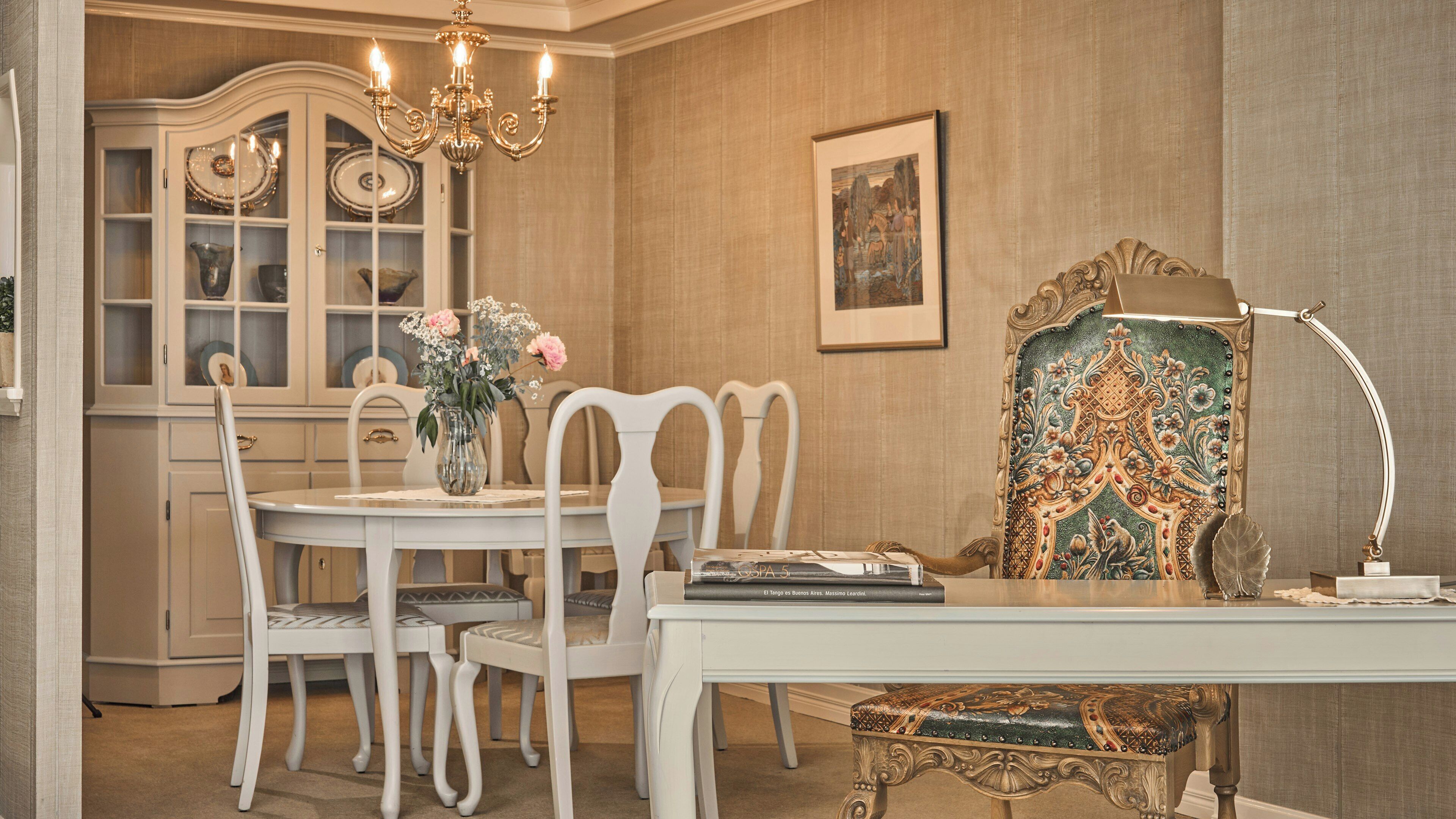 A dining area with a white table, chairs, a vase with flowers, a chandelier, a cabinet with glass doors, a framed painting, and a decorative lamp.