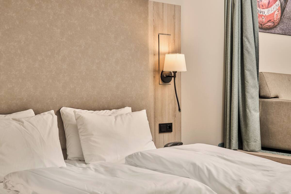 A hotel room with a bed, a wall-mounted lamp, a telephone, and a window with curtains.