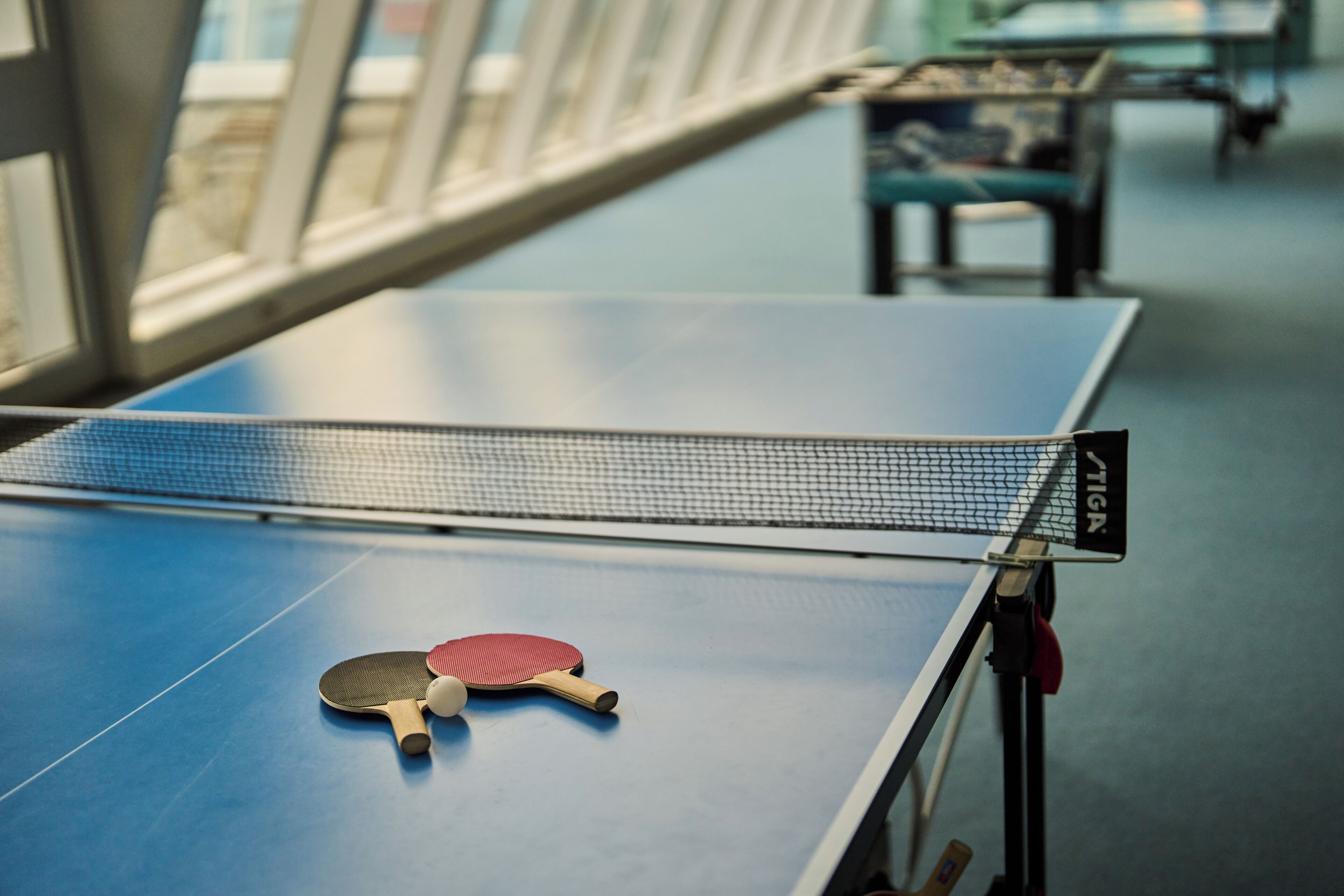 A ping pong table with two paddles and a ball, a net, and a chair in the background.