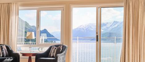 A room with a large sliding glass door leading to a balcony with a mountain view. There are two armchairs and a small table with a lamp.