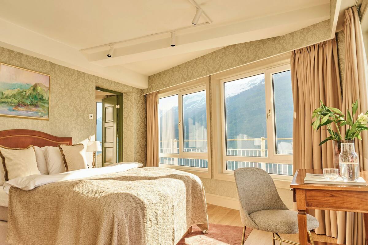 A hotel room with a large bed, a desk, a chair, a painting, and a view of mountains through the windows.