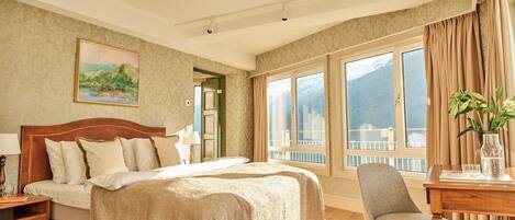 A hotel room with a large bed, a desk, a chair, a painting, and a view of mountains through the windows.