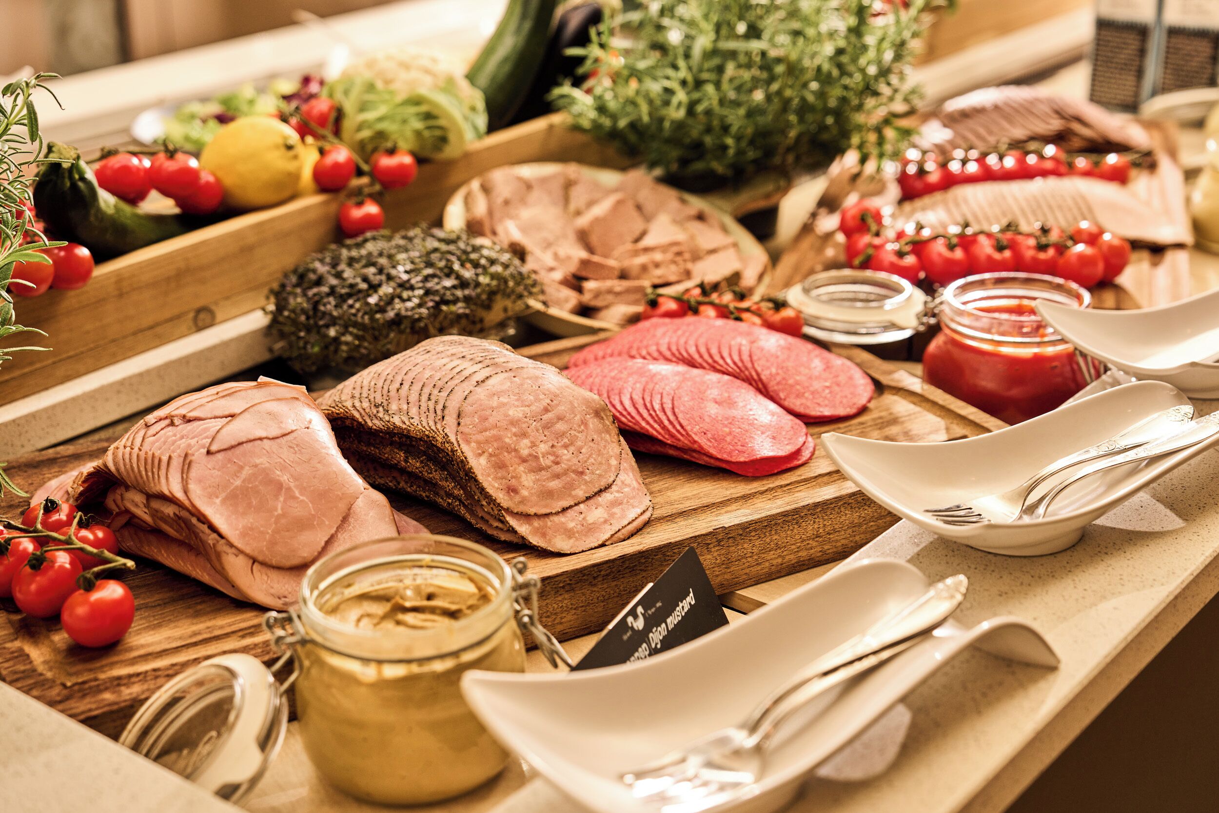 Variedad de carnes y verduras en una tabla de madera, incluyendo jamón en rodajas, tomates y un frasco de mostaza.
