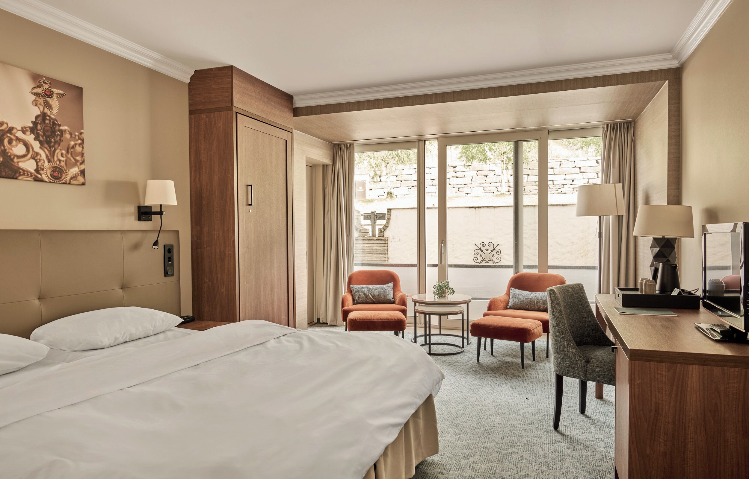A hotel room with a bed, two armchairs, a desk, a TV, and a large window with curtains.