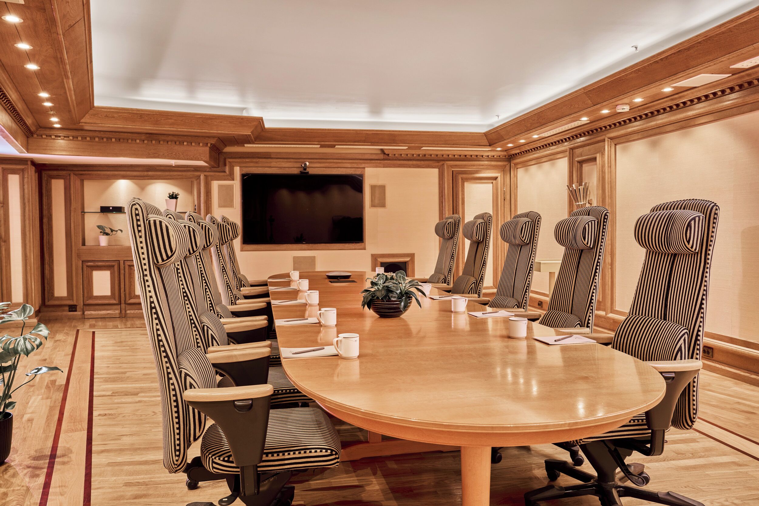 A conference room with a large oval table, high-backed chairs, a flat-screen TV, and a potted plant.