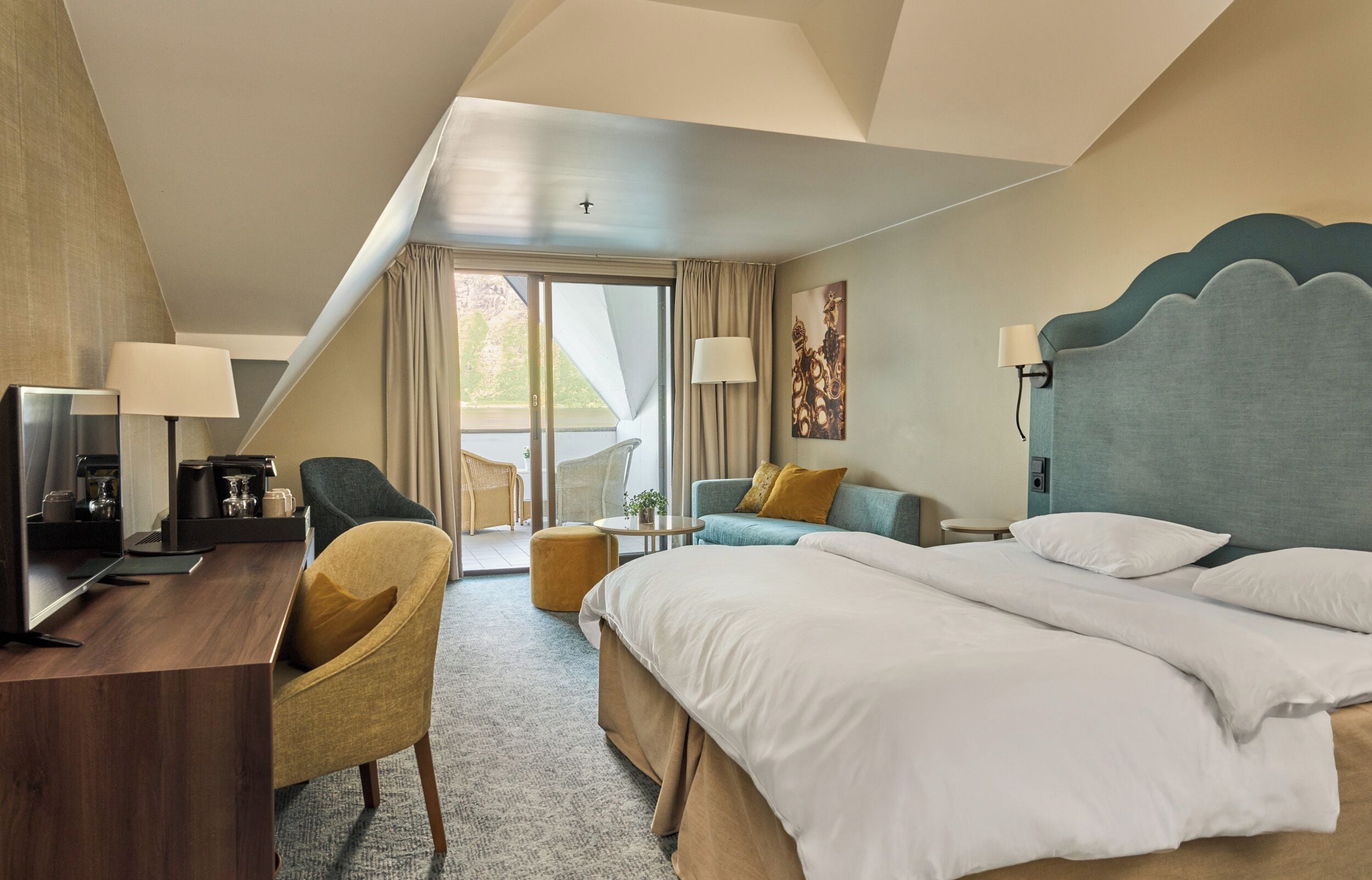 Quarto de hotel com cama, escrivaninha com computador, poltrona, sofá e varanda com cortinas.