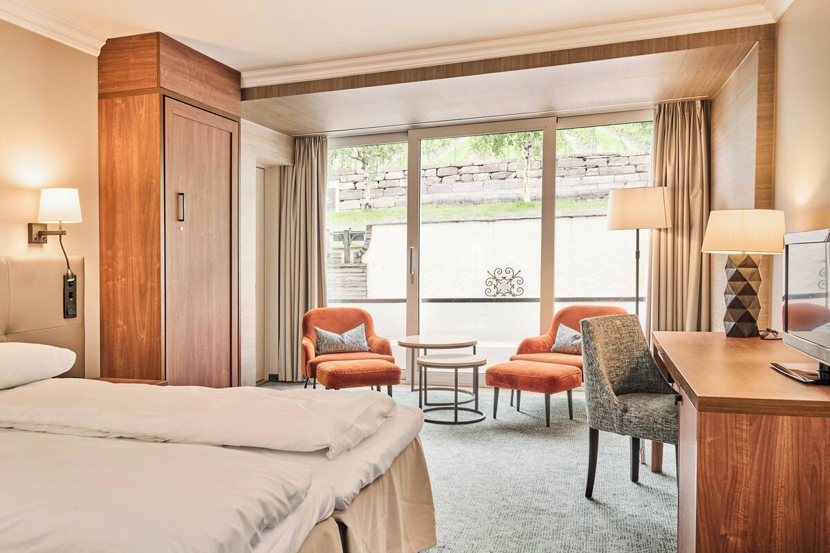 A hotel room with a bed, two armchairs, a desk with a TV, a small table, and a large window with curtains.