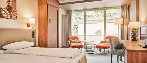 Habitación de hotel con cama, dos sillones, un escritorio con televisor, una mesita y un ventanal con cortinas.