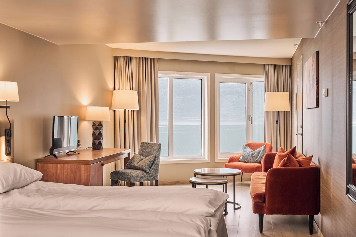 A hotel room with a bed, desk, chair, sofa, and large windows offering a view.