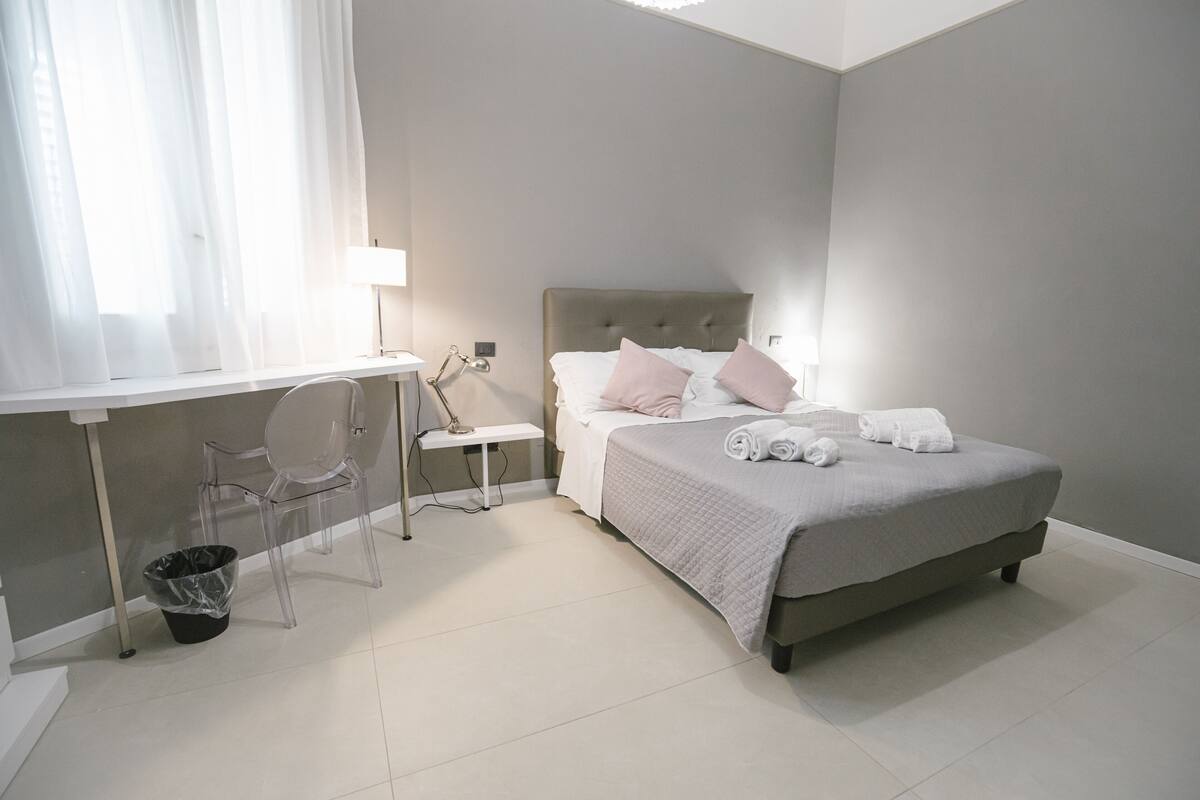 A bedroom with a bed, bedside table, lamp, chair, and a trash bin.