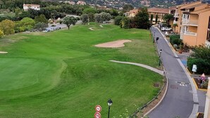 A golf course with a road curving alongside, surrounded by residential buildings and mountains in the background.